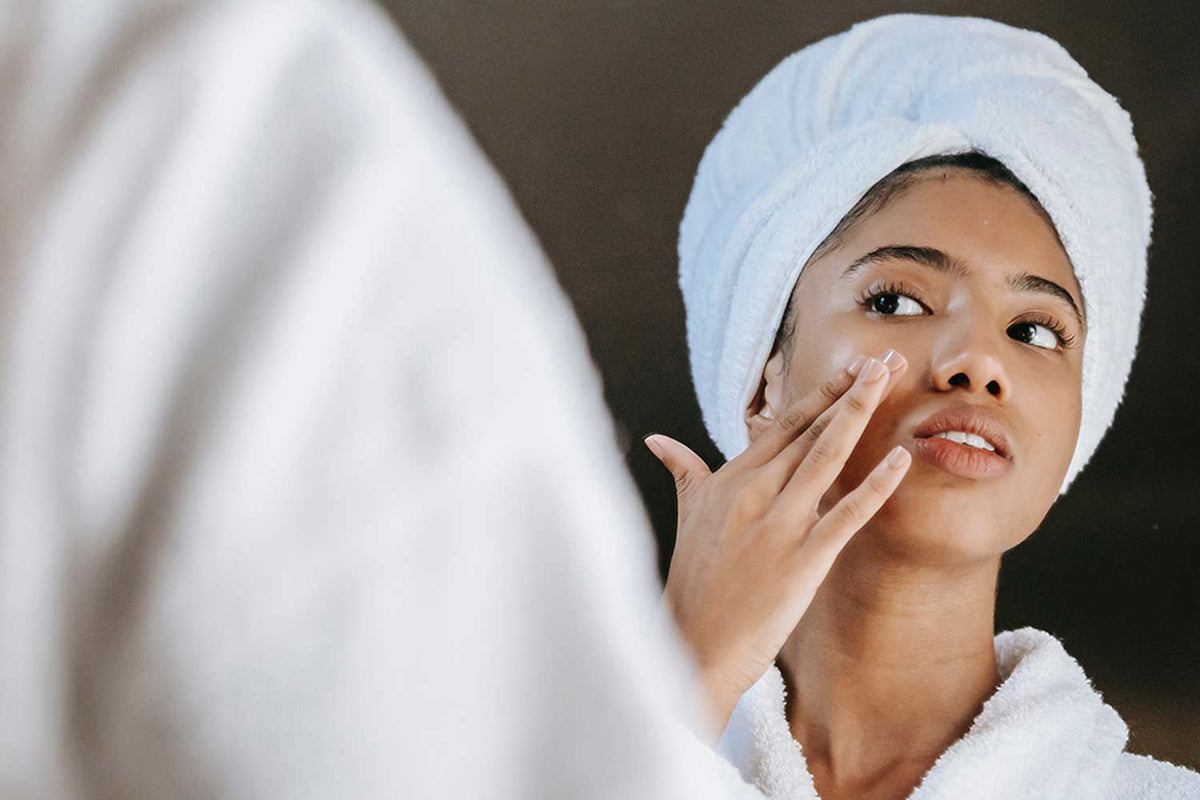 Lady looking into a mirror with a white hair towel on head applying skincare