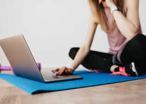lady in workout wear sitting on a yoga mat using a laptop